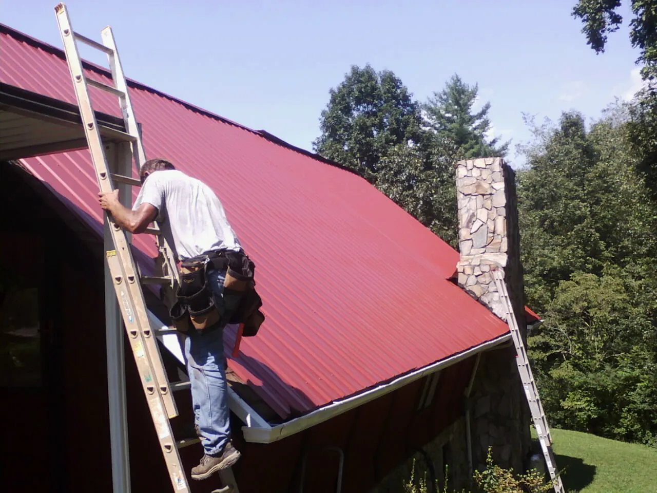 Roofer on a ladder beside a red metal roof and stone chimney — Soffit & Fascia Repair by Roofing Brothers in Moncks Corner